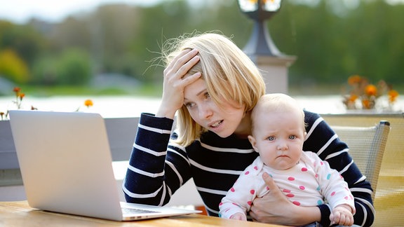 Eine verzweifelte alleinerziehende Mutter mit Baby auf dem Schoß vor dem Laptop, 
