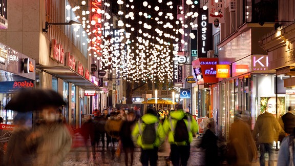 Passanten laufen mit Masken durch eine Einkaufsstrasse in der Köln, wo Weihnachtsbeleuchtung und Weihnachtsdekoration hängen. 