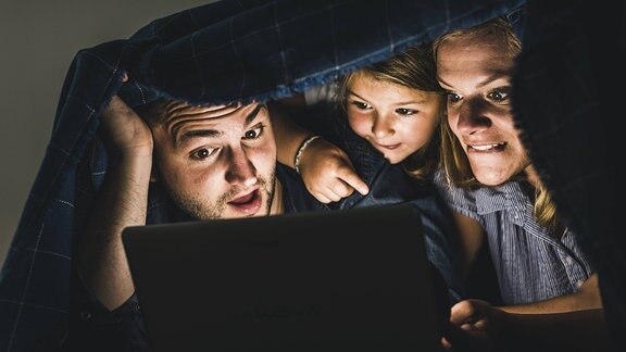 Eine Familie schaut auf dem Tablet gemeinsam einen Film. Sie haben eine Decke über ihre Kopfe und schauen gespannt auf den Bildschirm.