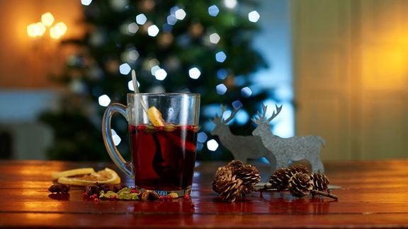 Glühwein in einer Glastasse auf einem Tisch im Vordergrund. Im Hintergrund leicht verschwommen ein Weihnachtsbaum.