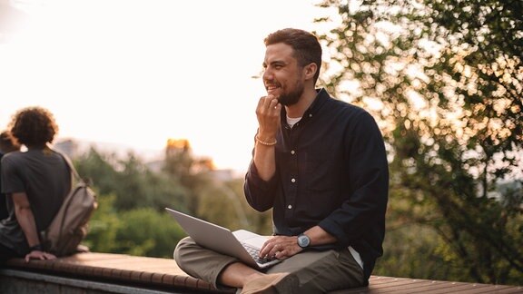 Fröhlicher nachdenklicher Mann mit Laptop, der auf einer Bank im Park sitzt