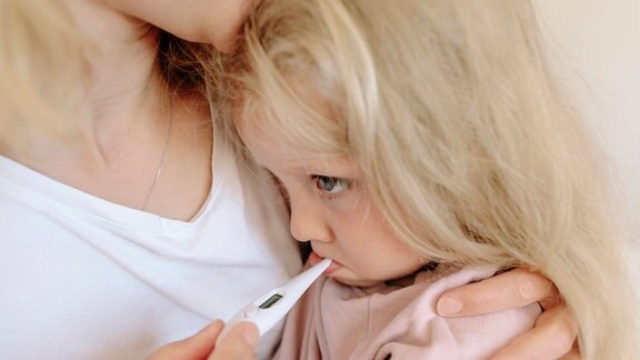 Mutter trägt Tochter und misst bei ihr Fieber mit einem Thermometer (Symbolfoto)