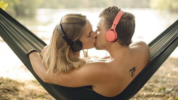 Ein Paar sitzt in der Hängematte und küsst sich. Beide haben Kopfhörer auf und hören Musik.