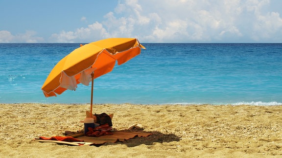 Ein orange-farbener Sonnenschirm steht an einem einsamen Strand, dahinter das blaue Meer.