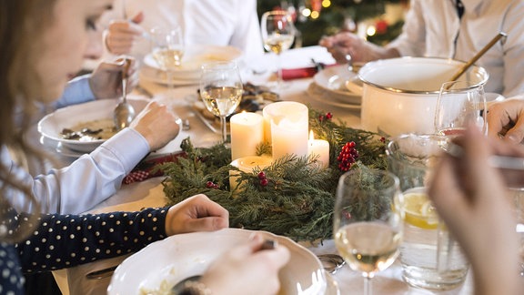 Familie sitzt um eine festliche Tafel zu Weihnachten. Es wird Suppe gegessen, Weingläser stehen auf dem Tisch und ein Adventskranz mit brennenden Kerzen.